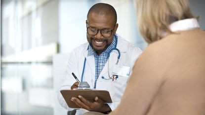 A doctor smiles as he writes on notepad while speaking with an older female patient.