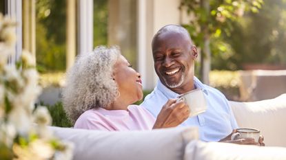 An older couple smile at each other as they enjoy coffee on their porch.