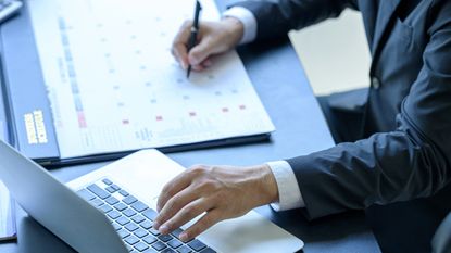 A businessman looks at a calendar while also using his laptop at his desk.