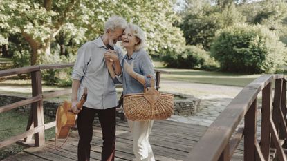 An older man holding a guitar and an older woman with a picnic basket laugh and hold hands in a park.