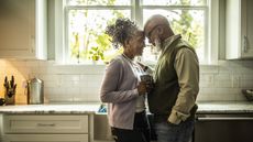 An older couple stand closely together in the kitchen, looking lovingly at each other.