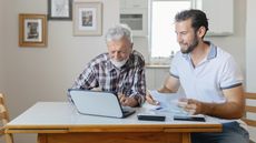 An adult son helps his father with his finances while sitting at the dining room table.