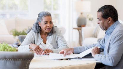 An older couple check over paperwork while sitting at their dining room table.