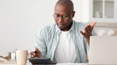 A man looks at his calculator on his desk and makes a "why is that number so high?" gesture.